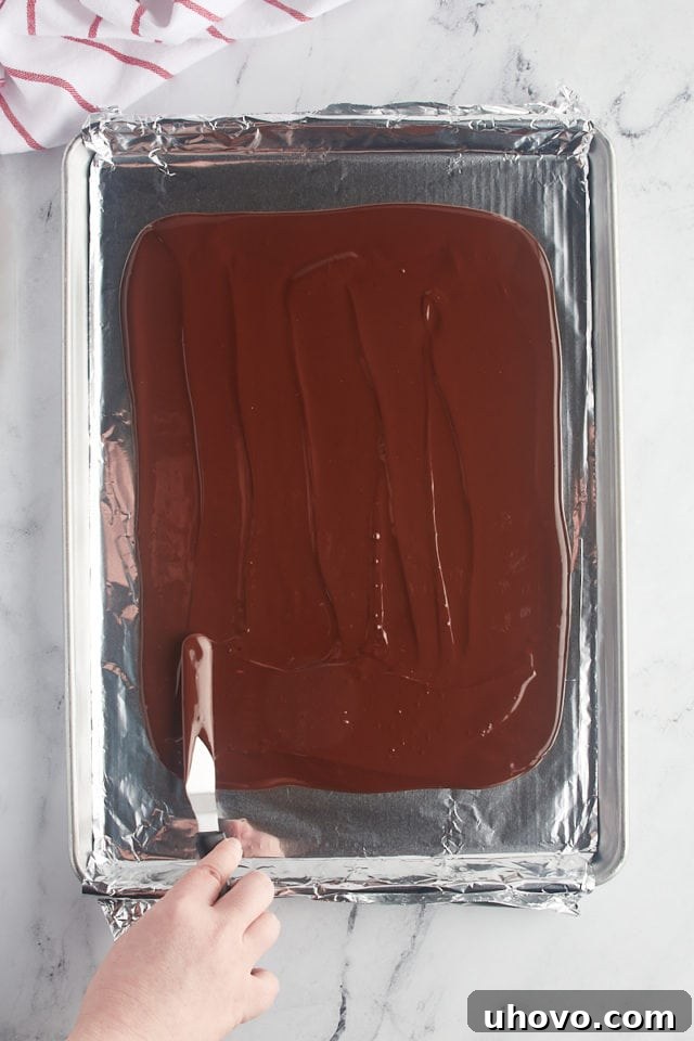 Overhead view of melted dark chocolate being carefully spread into an even layer on a foil-lined baking pan.