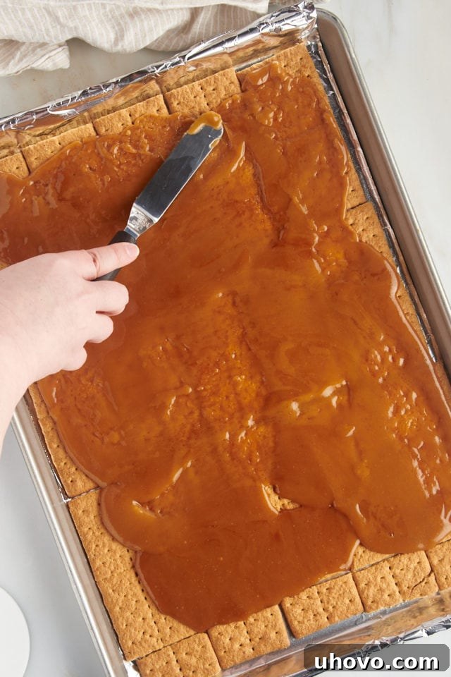 Overhead view of the warm caramel mixture being evenly spread over the graham cracker base in a half sheet pan.