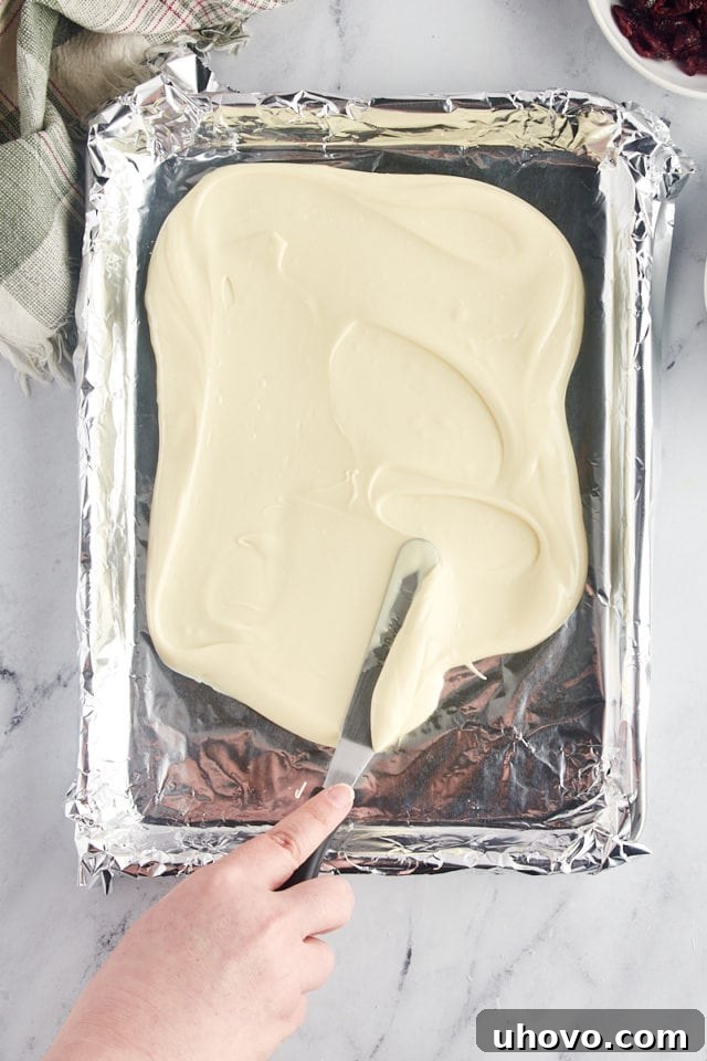 overhead view of melted white chocolate being smoothly spread into an even layer on a foil-lined baking sheet using an offset spatula