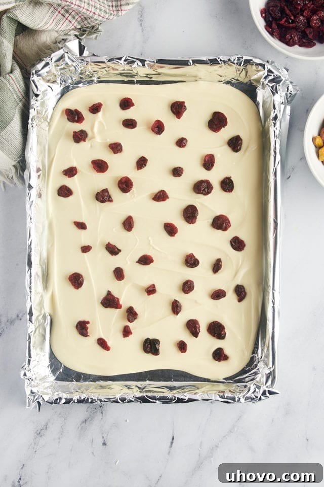 overhead view of bright red dried cranberries and green pistachios generously scattered over the still-wet melted white chocolate on a foil-lined baking sheet