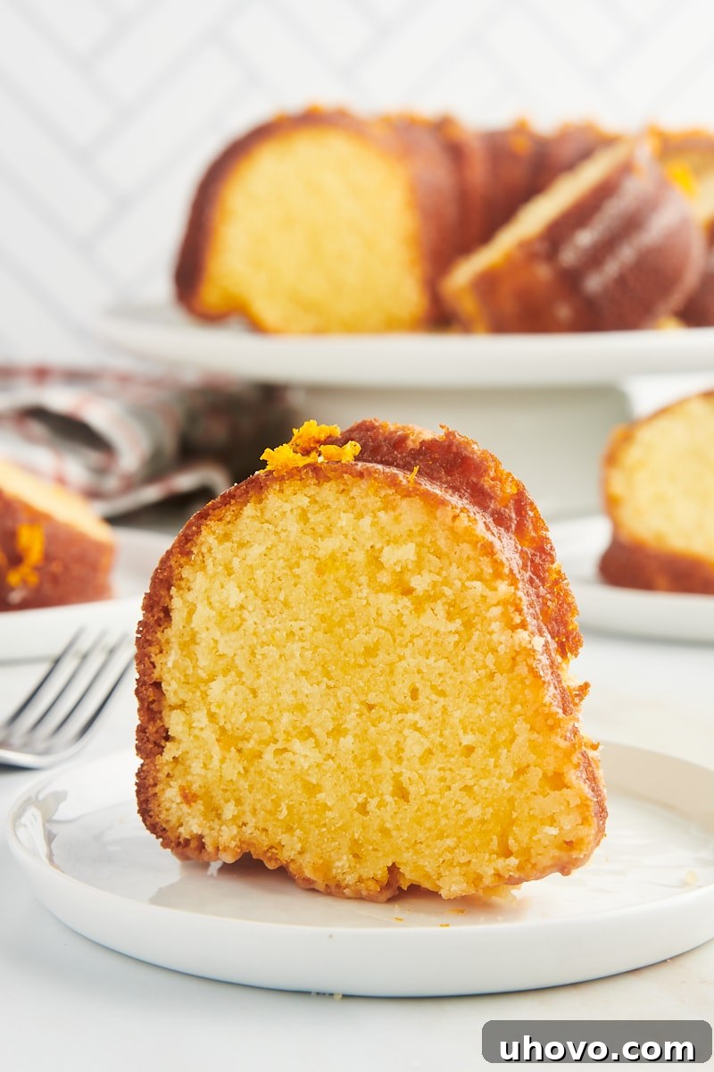 A single, delectable slice of orange pound cake on a white plate, with a larger cake on a stand blurred in the background, inviting a second serving.