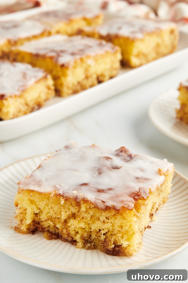 A perfectly portioned slice of honey bun cake on a white plate, with the rest of the cake slightly blurred in the background, inviting viewers to savor the moment.