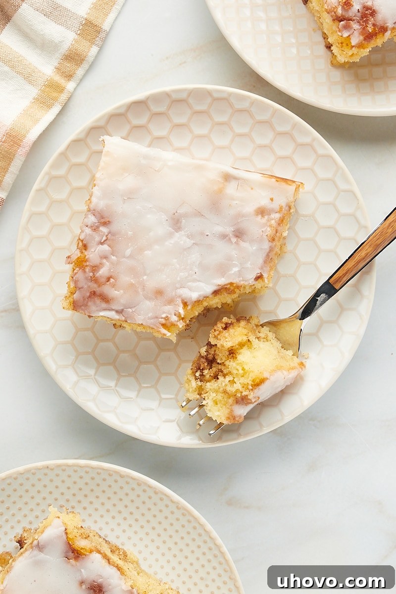 A close-up shot of a single slice of honey bun cake on a plate with a fork, emphasizing its moist interior, cinnamon swirl, and thick vanilla glaze, ready to be enjoyed.