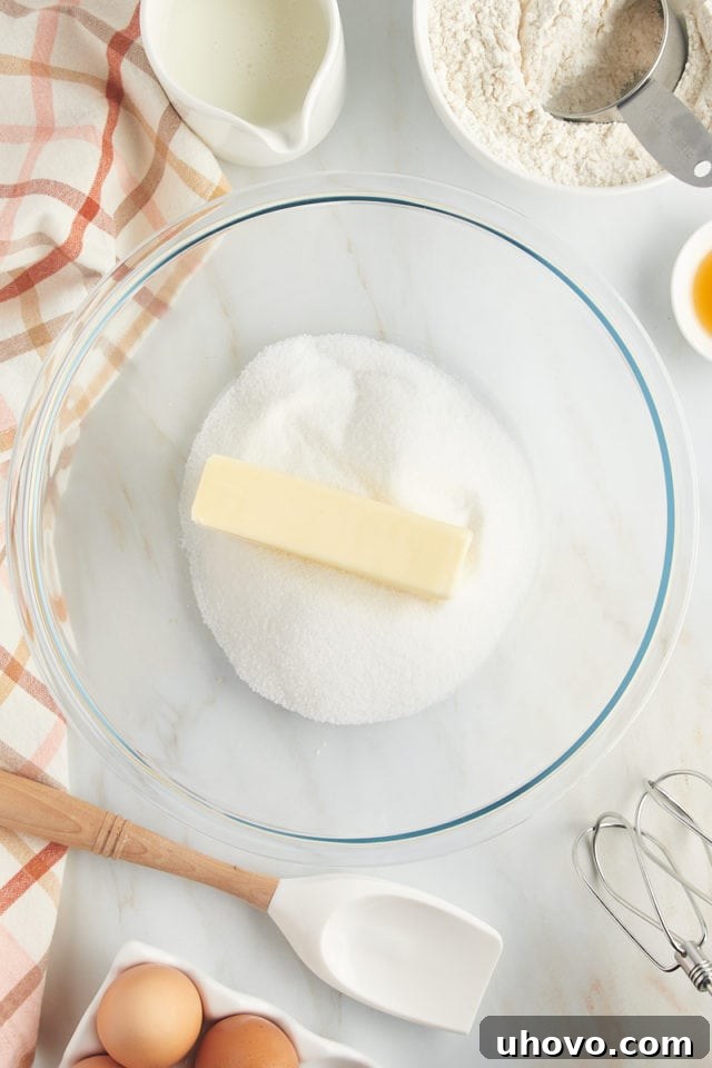An aerial view showing a stick of softened unsalted butter being added to a bowl of granulated sugar, ready for creaming to create a fluffy cake base.