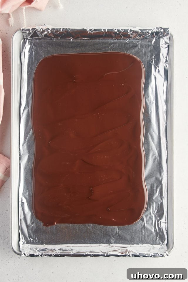 Overhead view of melted dark chocolate spread evenly on a foil-lined half sheet pan, forming the base of the bark.