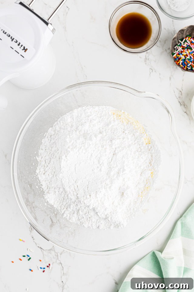 Overhead view of sifted confectioners' sugar being gradually added to the creamed butter in a glass mixing bowl for vanilla buttercream.