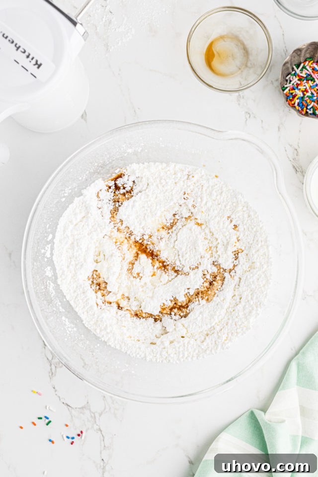 Overhead view of vanilla extract and milk being streamed into the frosting mixture in a glass mixing bowl, to achieve a smooth consistency.