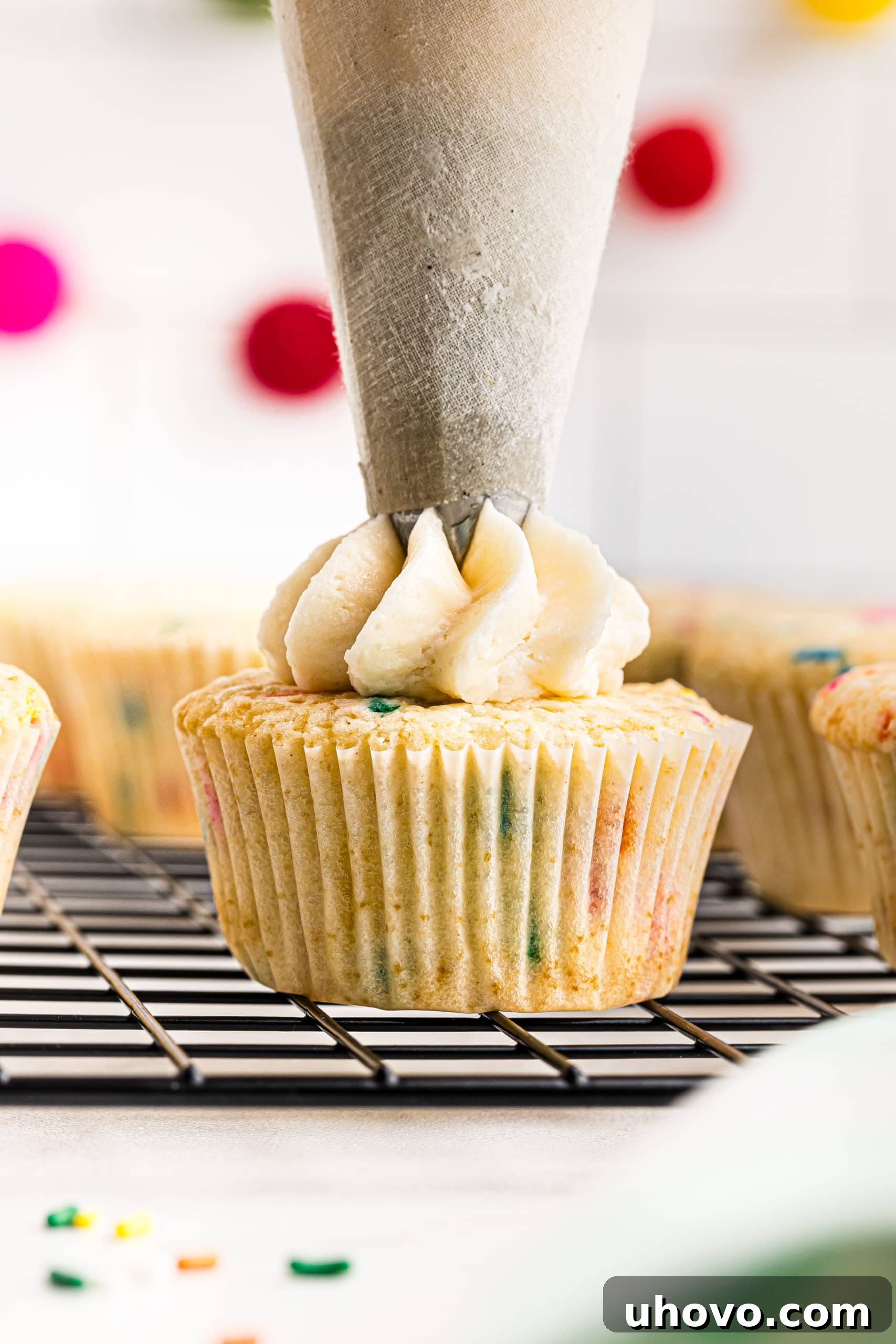 Smooth vanilla buttercream is expertly piped onto a single confetti cupcake, which rests on a wire rack, ready for its final festive touch.