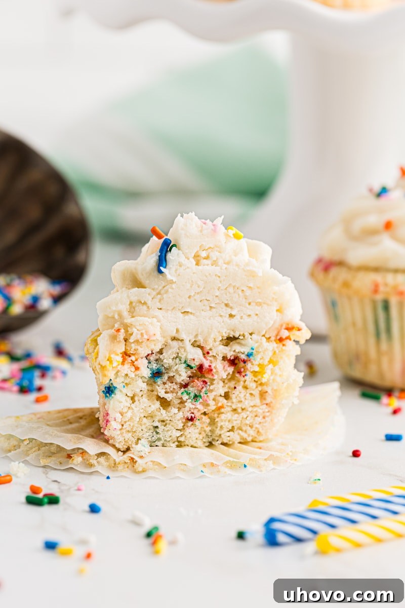 A perfectly halved confetti cupcake reveals its colorful sprinkle-filled interior, with another whole cupcake and loose sprinkles visible in the soft background.