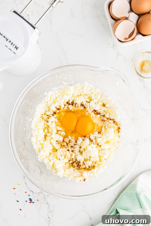 Overhead view of large eggs and vanilla extract being added to the creamed butter and sugar mixture in a glass mixing bowl, for confetti cupcakes.