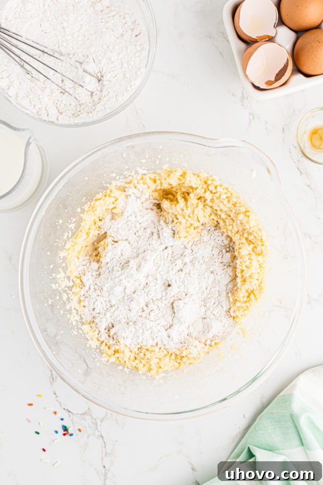 Overhead view of dry ingredients (flour mixture) being gently added to the wet cake batter in a glass mixing bowl, for confetti cupcakes.