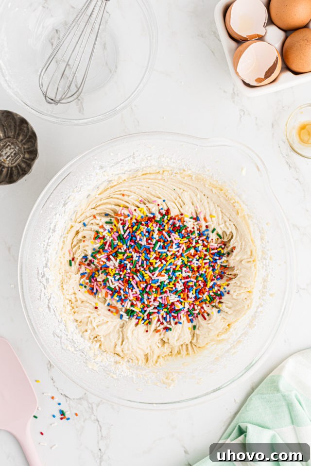 Overhead view of colorful jimmie sprinkles being gently folded into the vanilla cupcake batter in a glass mixing bowl, creating confetti cupcakes.