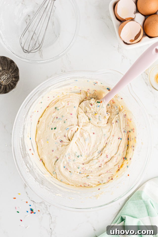 Overhead view of the finished confetti cupcake batter, speckled with vibrant sprinkles, resting in a glass mixing bowl, ready for baking.