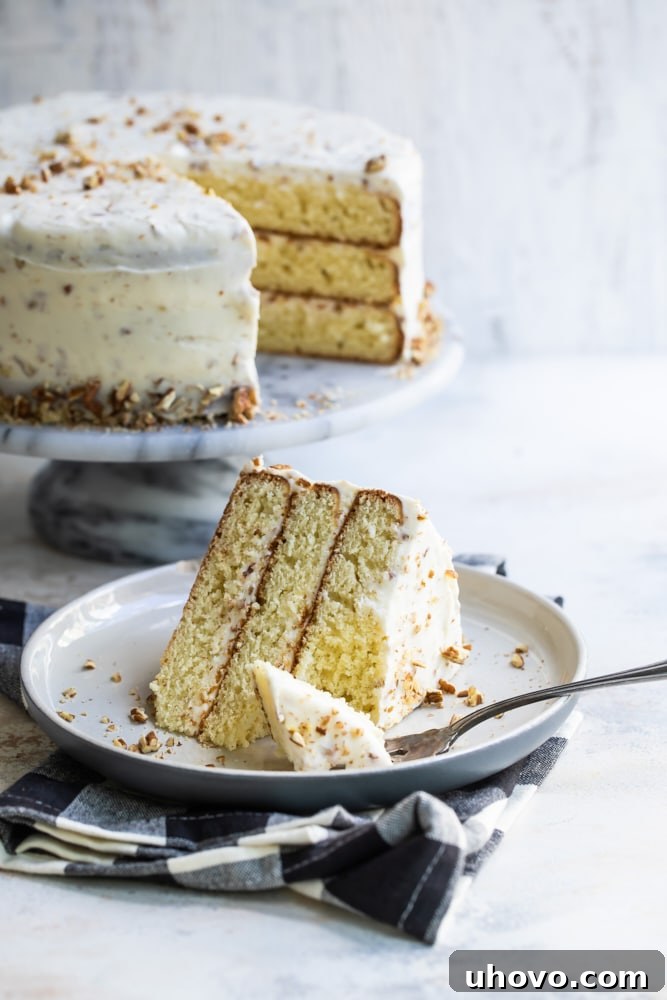 A delicious slice of Butter Pecan Cake on a beige dessert plate, with the remainder of the cake in the background on a marble cake stand.