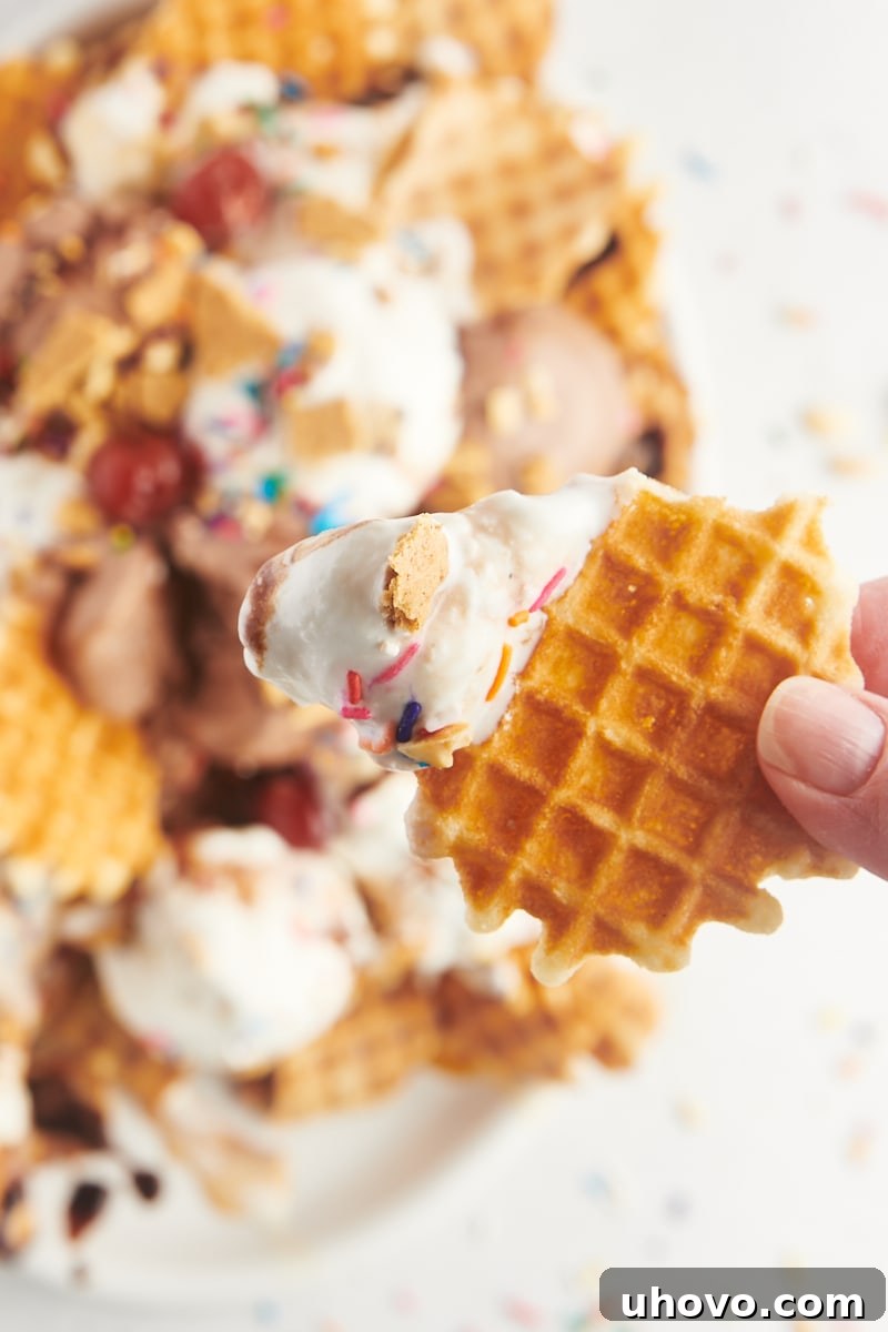 A hand holds a single broken waffle cone chip, delicately scooped with vanilla ice cream, topped with a drizzle of chocolate sauce and colorful sprinkles, ready to be enjoyed.