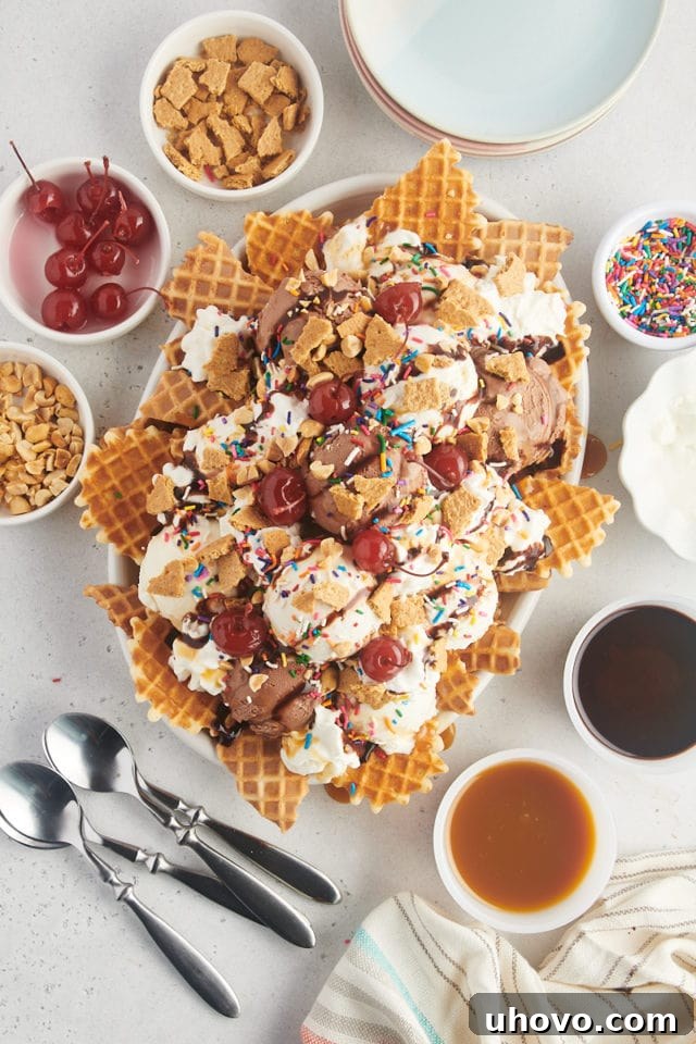 A vibrant, finished platter of ice cream nachos, loaded with various ice cream flavors and a colorful assortment of toppings, surrounded by small bowls of additional toppings, plates, and spoons, ready for serving.