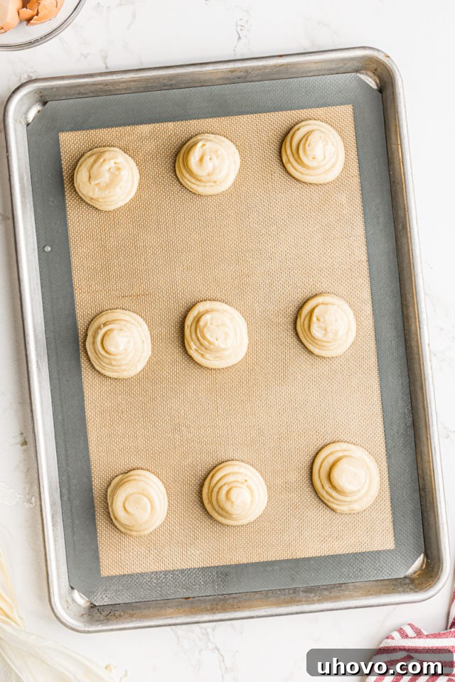 Overhead view of perfectly piped pâte à choux mounds arranged on a baking sheet lined with a silicone mat, ready for the oven.