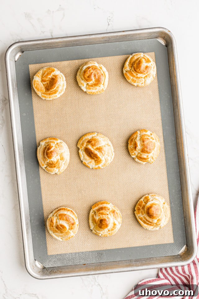 Overhead view of freshly baked, golden-brown cream puffs on a baking sheet lined with a silicone baking mat, having puffed up beautifully.