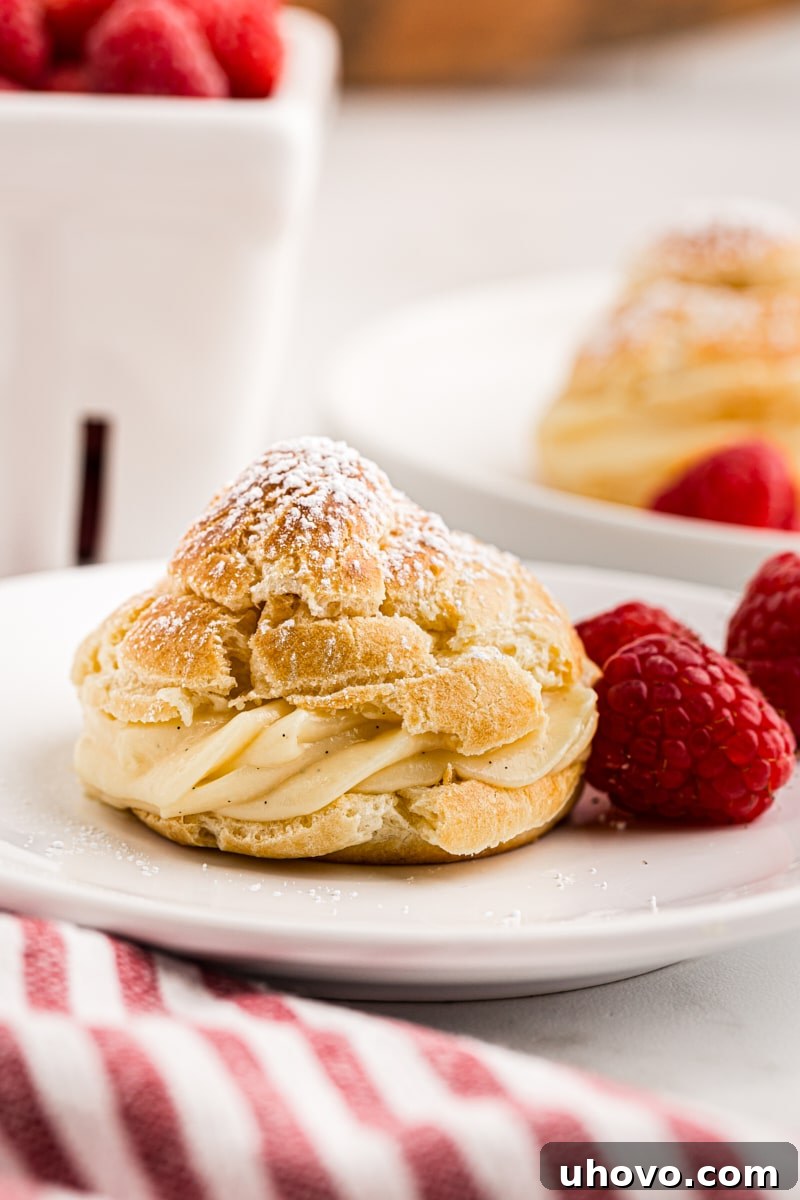 A cream puff and fresh raspberries on a white plate, with a container of raspberries and another cream puff blurred in the background, creating a delicious scene.