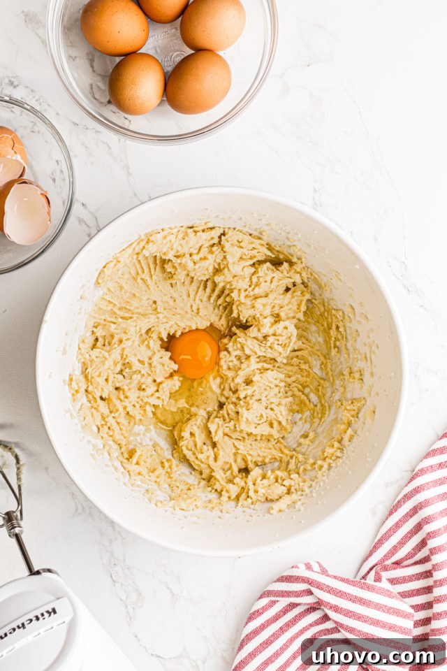 A single raw egg being cracked and added into the cooled pâte à choux dough in a mixing bowl, ready for incorporation.