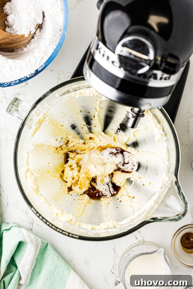 Overhead view of vanilla extract, salt, and half of the heavy cream being added to the lightened butter in a stand mixer bowl.
