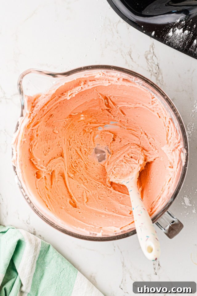 Overhead view of vibrant pink frosting in a mixing bowl, being pressed against the sides with a spatula to remove air bubbles.