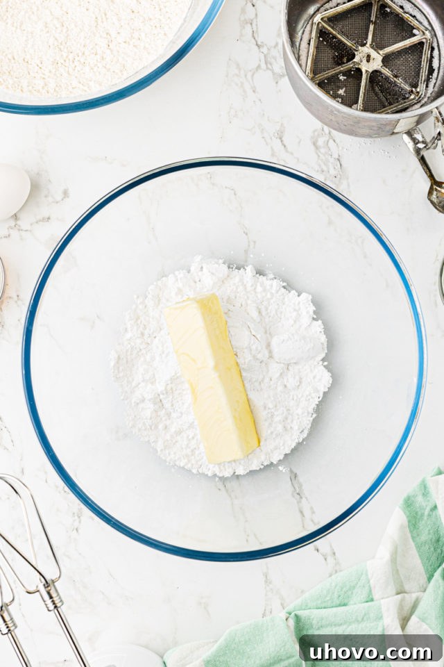 Overhead view of softened butter and confectioners' sugar in a clear glass mixing bowl, ready for creaming.
