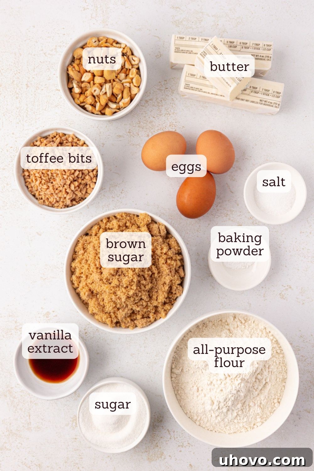 Overhead view of various ingredients for brown butter blondies laid out on a white surface, including butter, flour, sugars, eggs, vanilla, nuts, and toffee bits.