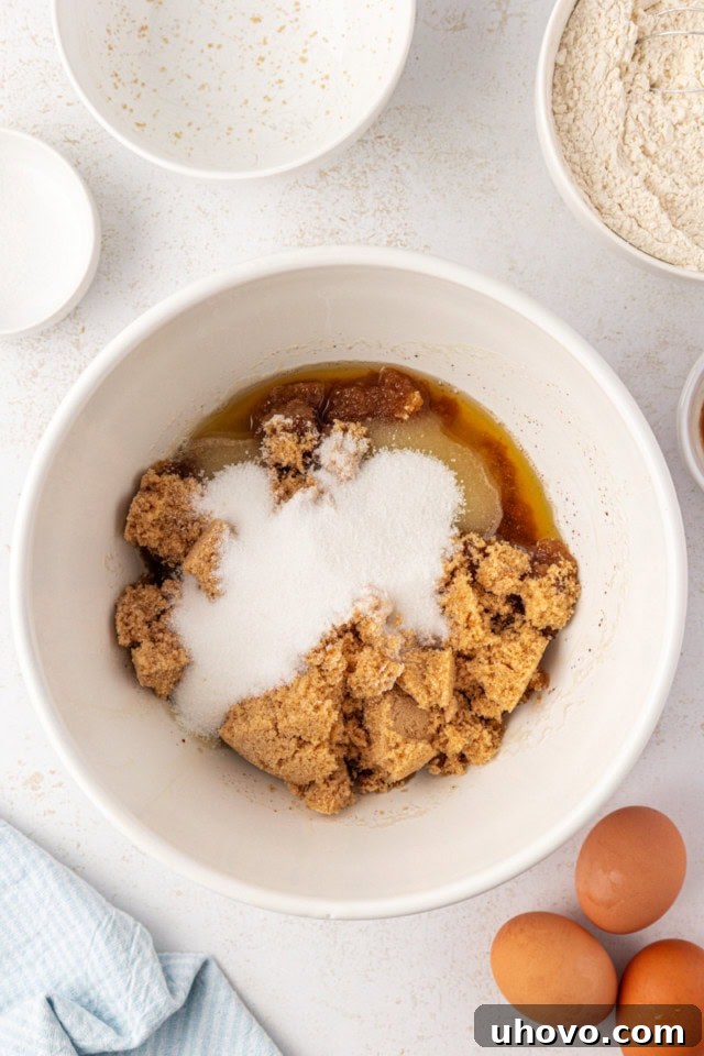 Overhead view of light brown sugar and granulated sugar being added to the slightly cooled brown butter in a large white mixing bowl.