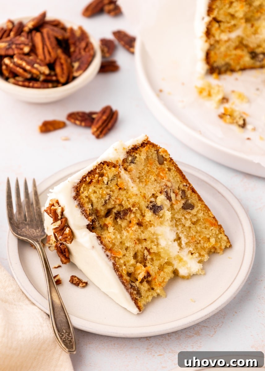 a slice of carrot cake on a light beige plate with more cake and a bowl of pecans in the background