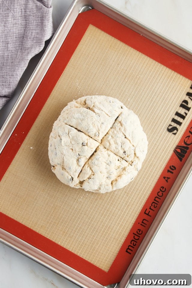 The raw Irish soda bread loaf, perfectly round and sitting on a baking sheet, has a deep 'X' scored across its top, indicating preparation for baking.