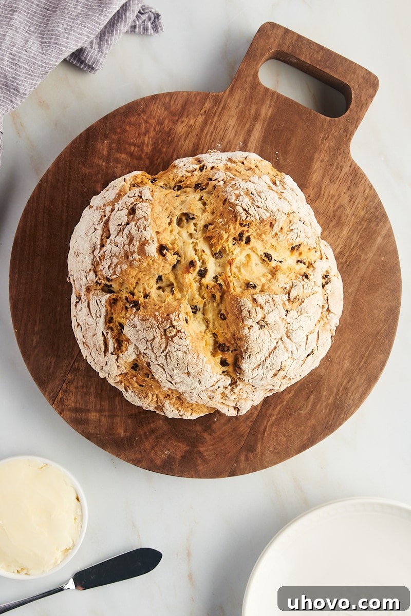 A bountiful Irish soda bread on a round wooden board, surrounded by a small dish of butter and stack of white serving plates.