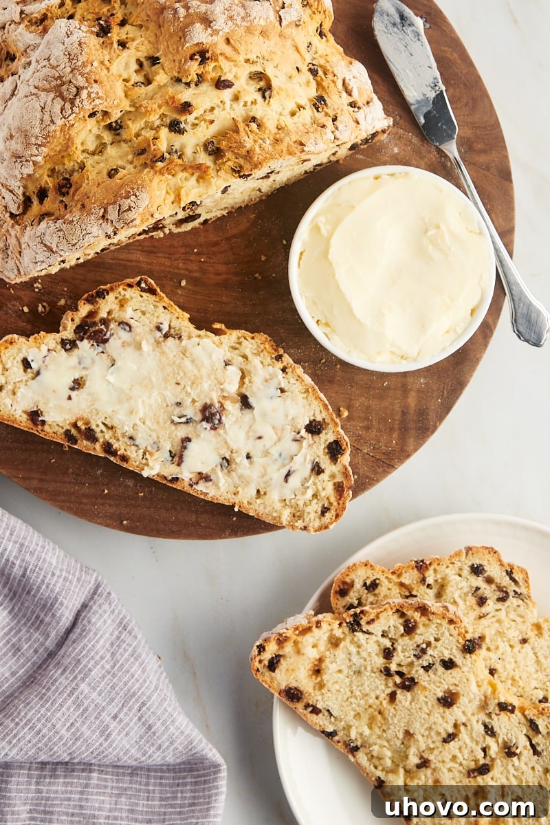 A close-up of a single slice of Irish soda bread, generously spread with butter, next to another warm loaf and a small dish of butter.