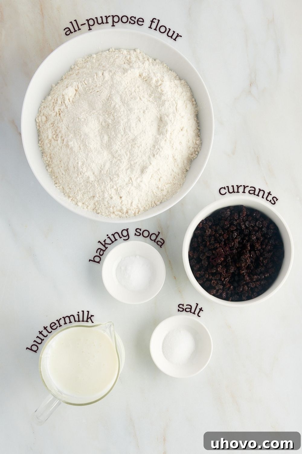 An overhead view showcasing the simple ingredients for Irish soda bread laid out on a clean counter: flour, baking soda, salt, buttermilk, and dried currants.