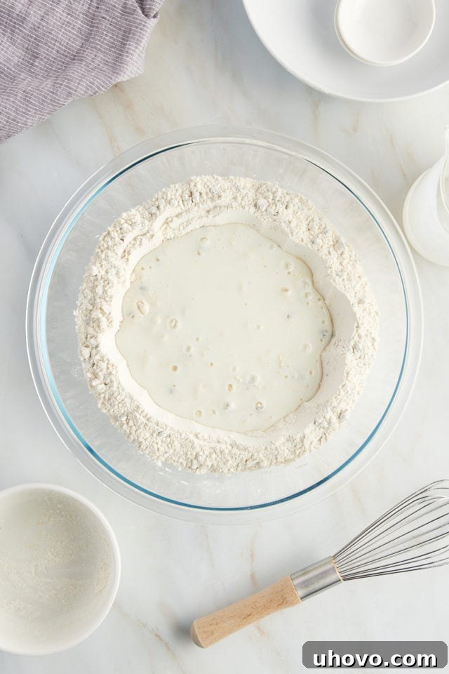 Creamy buttermilk is being poured precisely into the well of dry ingredients in a glass mixing bowl, initiating the dough formation.