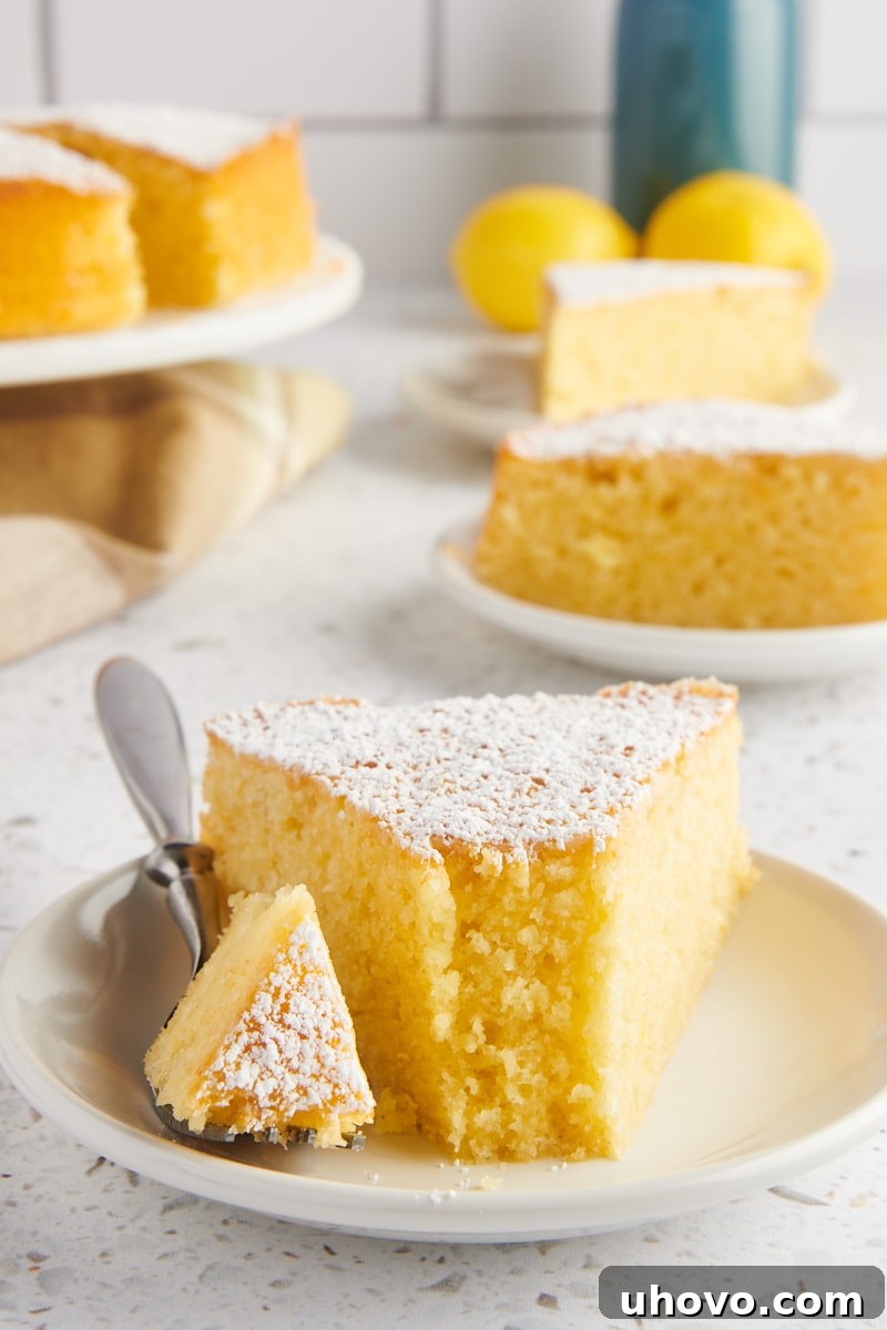 a slice of olive oil cake on a white plate with a bite on a fork; more cake and lemons in the background