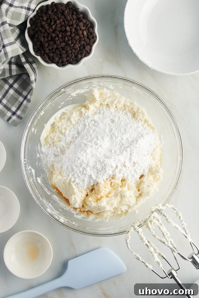 overhead view of confectioners' sugar, vanilla extract, and almond extract added to combined mascarpone and ricotta cheeses in a glass mixing bowl