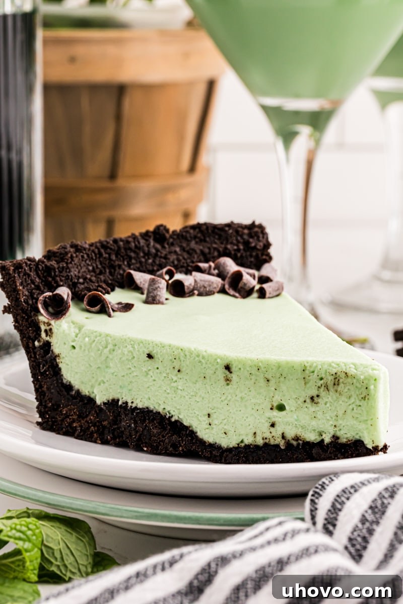 A vibrant slice of Grasshopper Pie on a white plate, garnished with chocolate shavings and a mint leaf, with a classic Grasshopper cocktail blurred in the background.