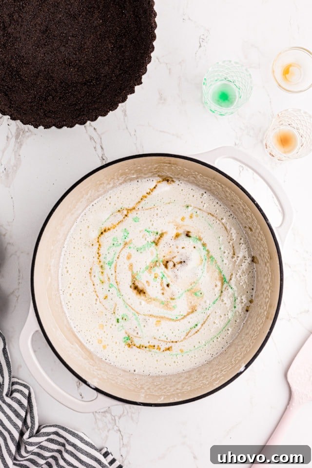 Crème de menthe, crème de cacao, and vanilla extract being stirred into the melted marshmallow mixture in a large saucepan, adding color and flavor.