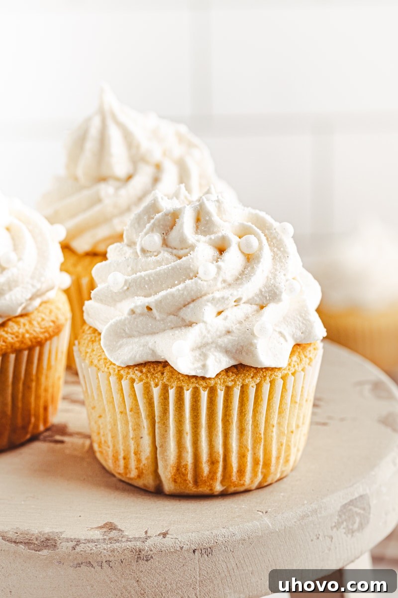 A perfectly frosted vanilla cupcake on a wooden cake stand with more blurred cupcakes in the background, showcasing its fluffy texture and elegant presentation.