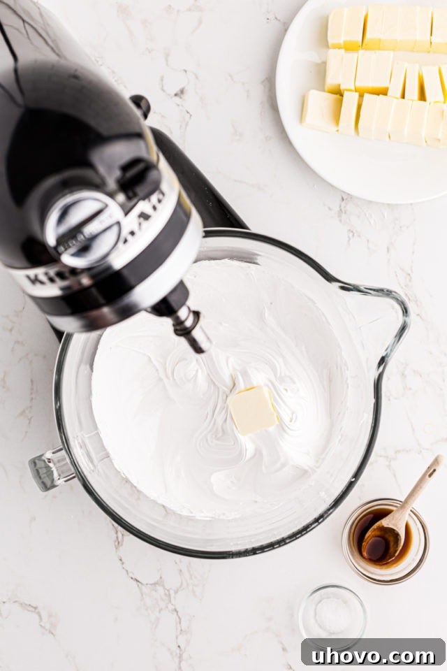 Overhead view of softened, cubed butter being gradually added to the whipped Swiss meringue in the bowl of a stand mixer.