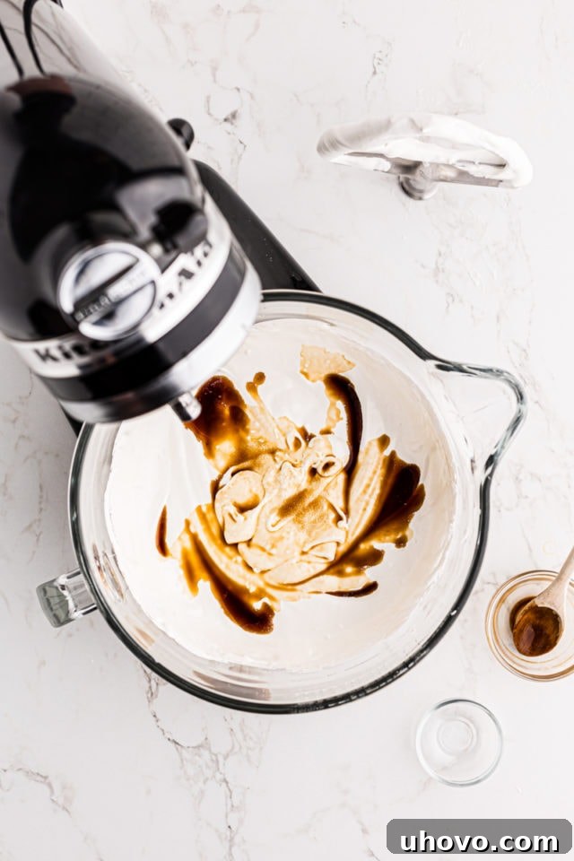 Overhead view of salt and vanilla extract being added to the Swiss meringue buttercream in the bowl of a stand mixer, ready for final mixing.