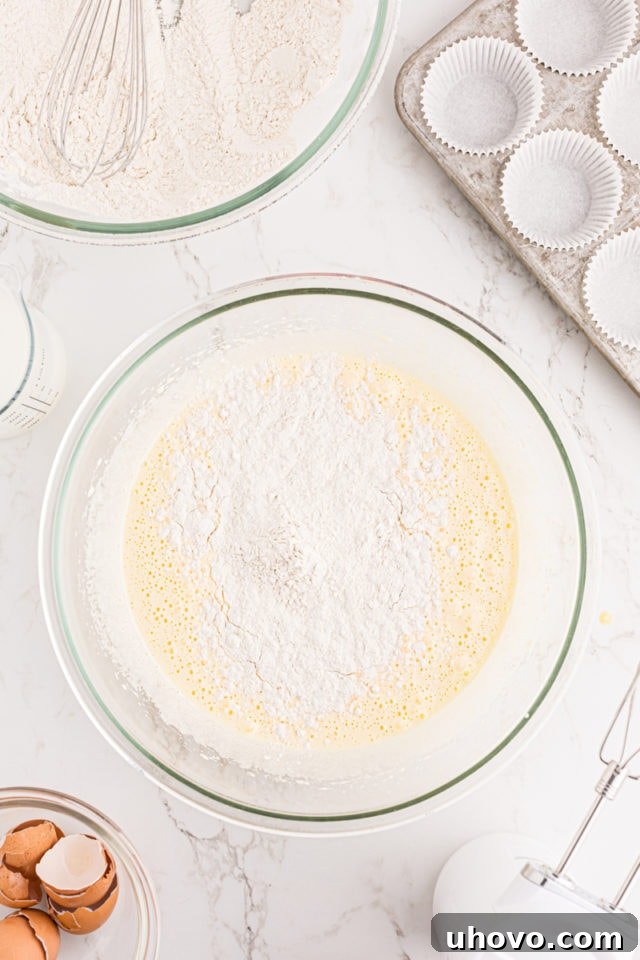 Overhead view of the dry flour mixture being gradually added to the wet vanilla cupcake batter in a glass mixing bowl, ready for gentle incorporation.