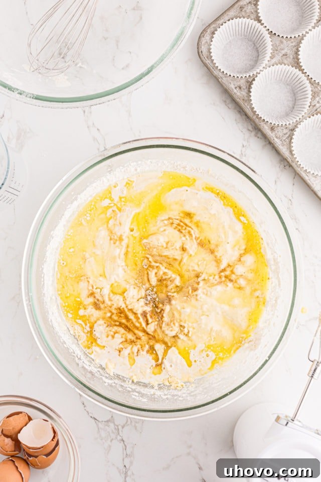 Overhead view of milk, vegetable oil, vanilla extract, and slightly cooled melted butter being poured into the vanilla cupcake batter in a glass mixing bowl.