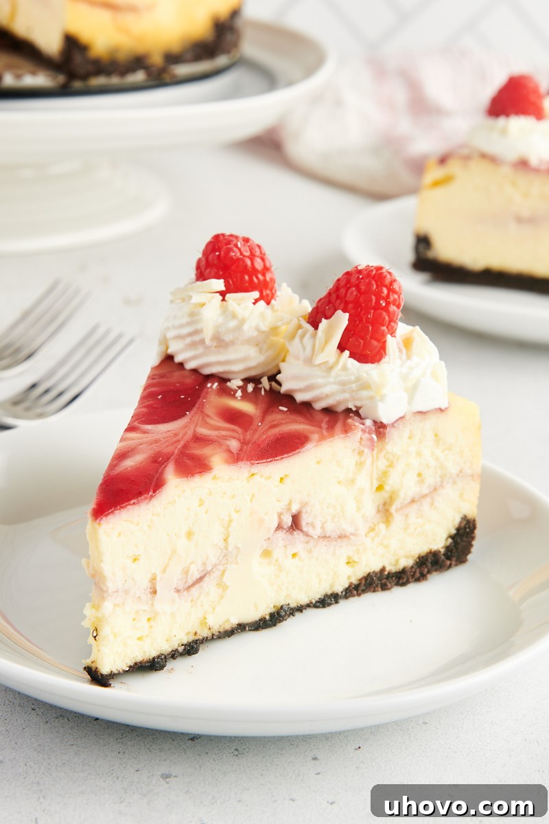 Side view of a generously sized white chocolate raspberry cheesecake slice on a plate, showing distinct layers of Oreo crust, creamy filling, and vibrant raspberry swirls, garnished with fresh raspberries and white chocolate shavings.