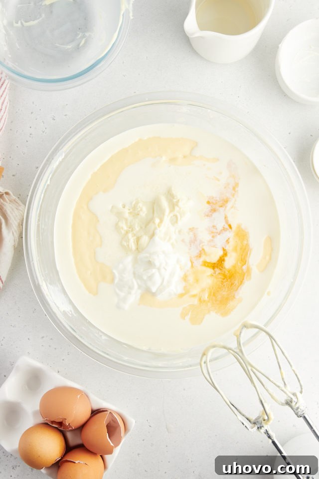 Overhead view of melted white chocolate, heavy cream, sour cream, vanilla extract, and almond extract added to the cream cheese mixture in a bowl.