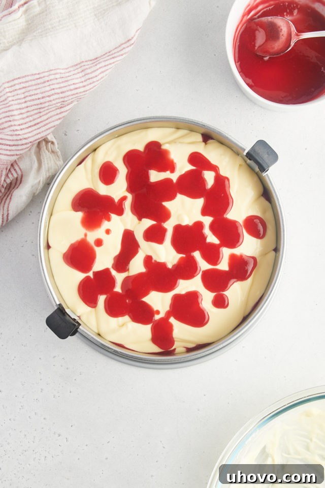 Overhead view of spoonfuls of vibrant raspberry sauce being artfully dolloped over the first layer of cheesecake filling, followed by the addition of the second layer of filling.