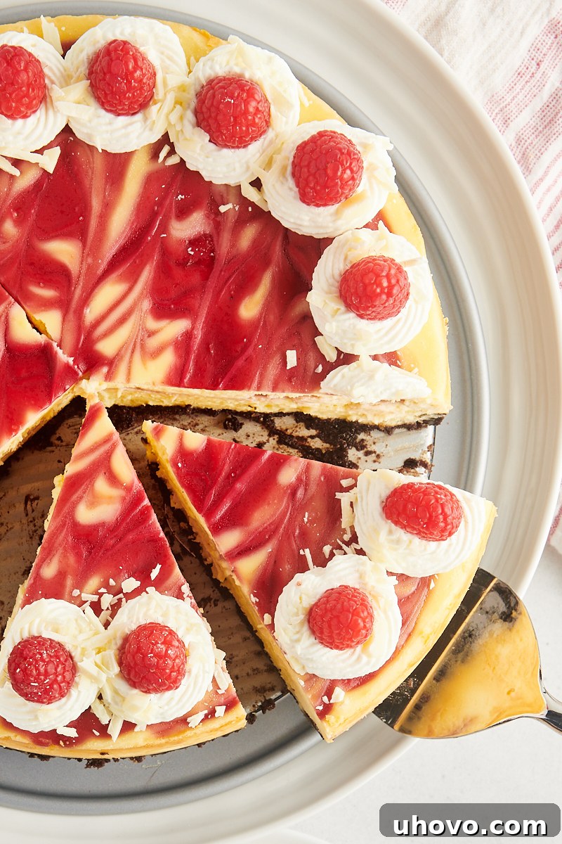 Overhead view of a beautifully cut white chocolate raspberry cheesecake presented on a round serving platter, showing perfect, clean slices with visible layers of Oreo crust, white chocolate filling, and distinct raspberry swirls.