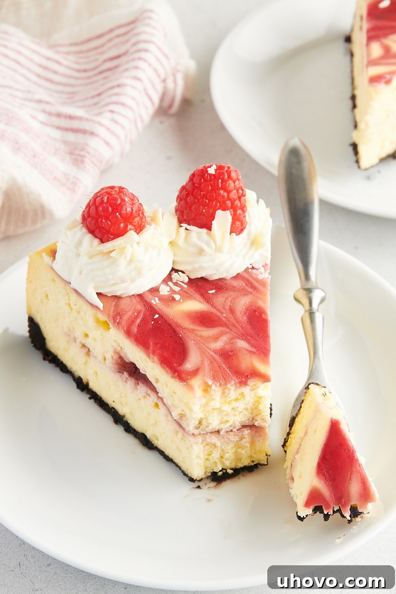 Close-up view of a perfect slice of white chocolate raspberry cheesecake on a white plate, with a fork gently taking a bite. The image highlights the smooth, creamy texture of the filling, the distinct raspberry swirl, and the sturdy Oreo crust.
