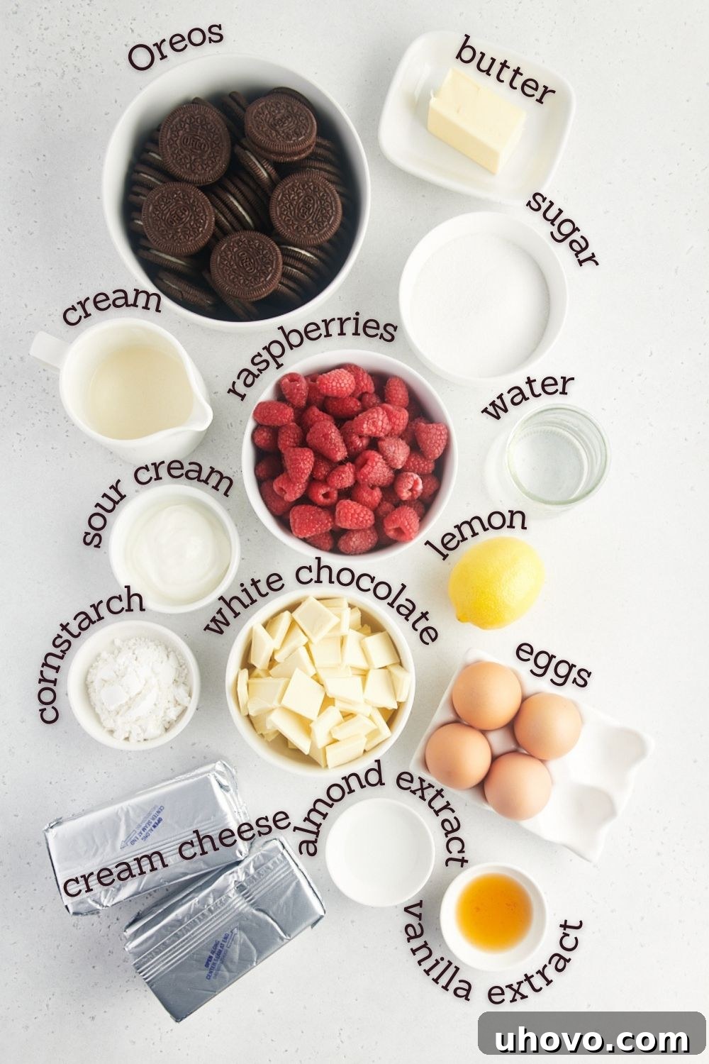 Overhead view of fresh ingredients laid out on a wooden surface for making white chocolate raspberry cheesecake, including blocks of cream cheese, fresh raspberries, white chocolate bars, and other baking essentials.